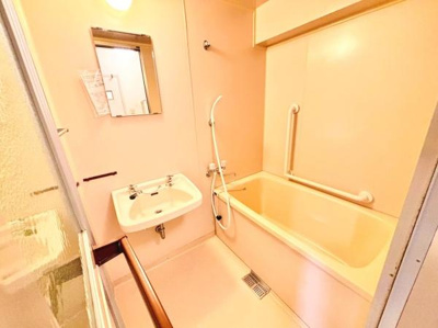 明るい色合いの洗面所と浴室で清潔感のある空間です。壁面には手すりが設置されており使いやすさも考慮されています。浴槽と洗面台の配置がコンパクトで効率的に利用できます。