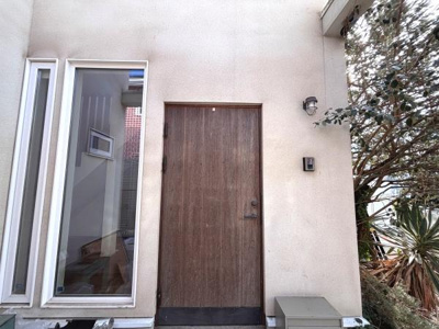 シンプルな木目調の扉が落ち着いた雰囲気を演出します。大きな縦長窓が隣接し自然光がしっかり差し込みます。玄関先には植栽もありさりげない緑がアクセントになっています。