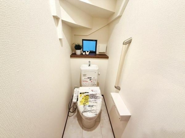 ホワイトを基調とした清潔感のあるトイレです。手すりが設置されており使いやすさが感じられます。収納スペースもあり小物を置くのに便利です。通路がしっかりと確保されていて移動もスムーズに行えます。