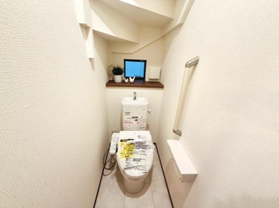 ホワイトを基調とした清潔感のあるトイレです。手すりが設置されており使いやすさが感じられます。収納スペースもあり小物を置くのに便利です。通路がしっかりと確保されていて移動もスムーズに行えます。