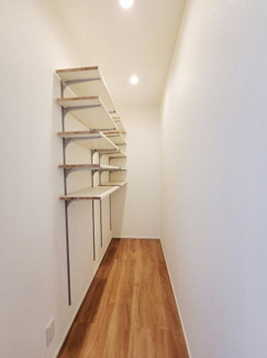 壁面に設置されたシンプルな棚が特徴の収納スペースです。縦長の空間で奥行きも確保されています。明るい照明で中のものが見やすい設計です。床の木目が空間に温かみを加えています。