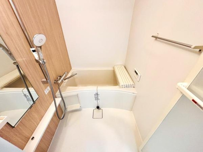 明るい色合いの内装が清潔感を感じさせる浴室です。壁面のウッド調パネルがアクセントになっています。シャワーや浴槽まわりも手入れしやすい設計です。タオル掛けや小物置きも備わっています。