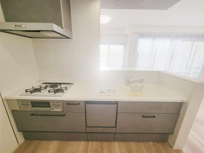 カウンター付きのキッチンは作業スペースが広めです。明るい室内が特徴的です。収納が多く整理しやすい設計です。床とキャビネットの色調が調和しています。ガスコンロや換気扇も設置されています。