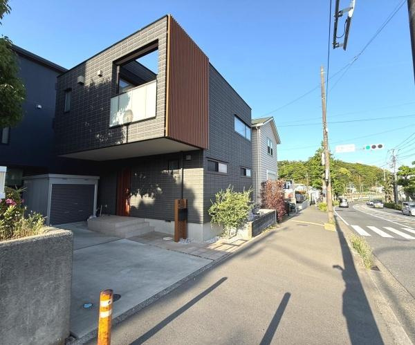 現地写真からは建物の外観がシンプルでスタイリッシュな印象を受けます。前面道路は広く歩道も整っています。角地に位置し車の出し入れもしやすそうです。周辺には緑も見られ落ち着いた環境が感じられます。