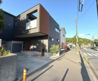 現地写真からは建物の外観がシンプルでスタイリッシュな印象を受けます。前面道路は広く歩道も整っています。角地に位置し車の出し入れもしやすそうです。周辺には緑も見られ落ち着いた環境が感じられます。