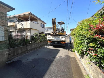 前面道路は舗装されており周辺には住宅が並んでいます。日差しが差し込み道路沿いの植栽も印象的です。現地の様子が分かりやすく周辺環境を確認できます。車両の進入も可能な道幅です。