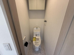シンプルで落ち着いたデザインのトイレです。上部には便利な収納棚が設置されています。壁紙や床材は明るいトーンで統一されています。
