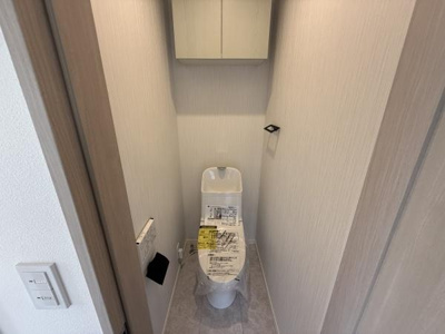 シンプルで落ち着いたデザインのトイレです。上部には便利な収納棚が設置されています。壁紙や床材は明るいトーンで統一されています。