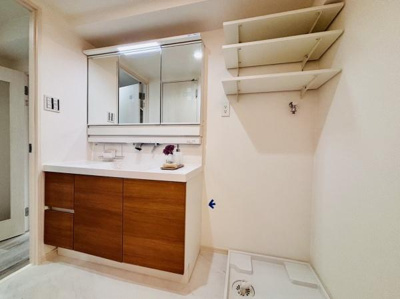 木目調の洗面台が空間に温かみを演出しています。大きな鏡付きキャビネットで毎日のお手入れが快適です。壁面には収納棚も設置されており使い勝手にも配慮されています。