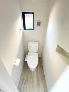 清潔感のある明るいトイレ空間です。窓付きで自然光が差し込み換気もしやすい仕様となっています。壁面の棚スペースも便利にご活用いただけます。木目調の床が落ち着いた雰囲気を演出しています。