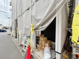 建物外観にはしっかりとした工事用の養生シートが設置されています。歩道沿いでアクセスしやすい立地です。現地の目印にはカラーコーンや脚立が設置されています。工事用資材も整理されています。