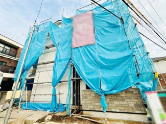 建築中の外観で新築ならではの清潔感が印象的です。シンプルなデザインが目を引きます。青いシートが現場の整然さを感じさせます。周囲の住環境にも調和しています。石調の外壁もアクセントとなっています。