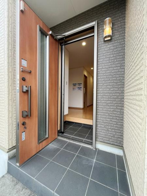 玄関ドアのデザインが印象的です。タイル貼りの床が落ち着いた雰囲気を演出しています。壁付けの照明器具が外観にアクセントを加えています。室内への動線もスムーズです。手入れのしやすい素材を採用しています。