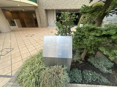 エントランス前の植栽が整えられており清潔感のある空間です。マンション名がしっかりと表示されたサインが設置されています。タイル張りの床が高級感を演出しています。緑の植え込みがアクセントになっています。